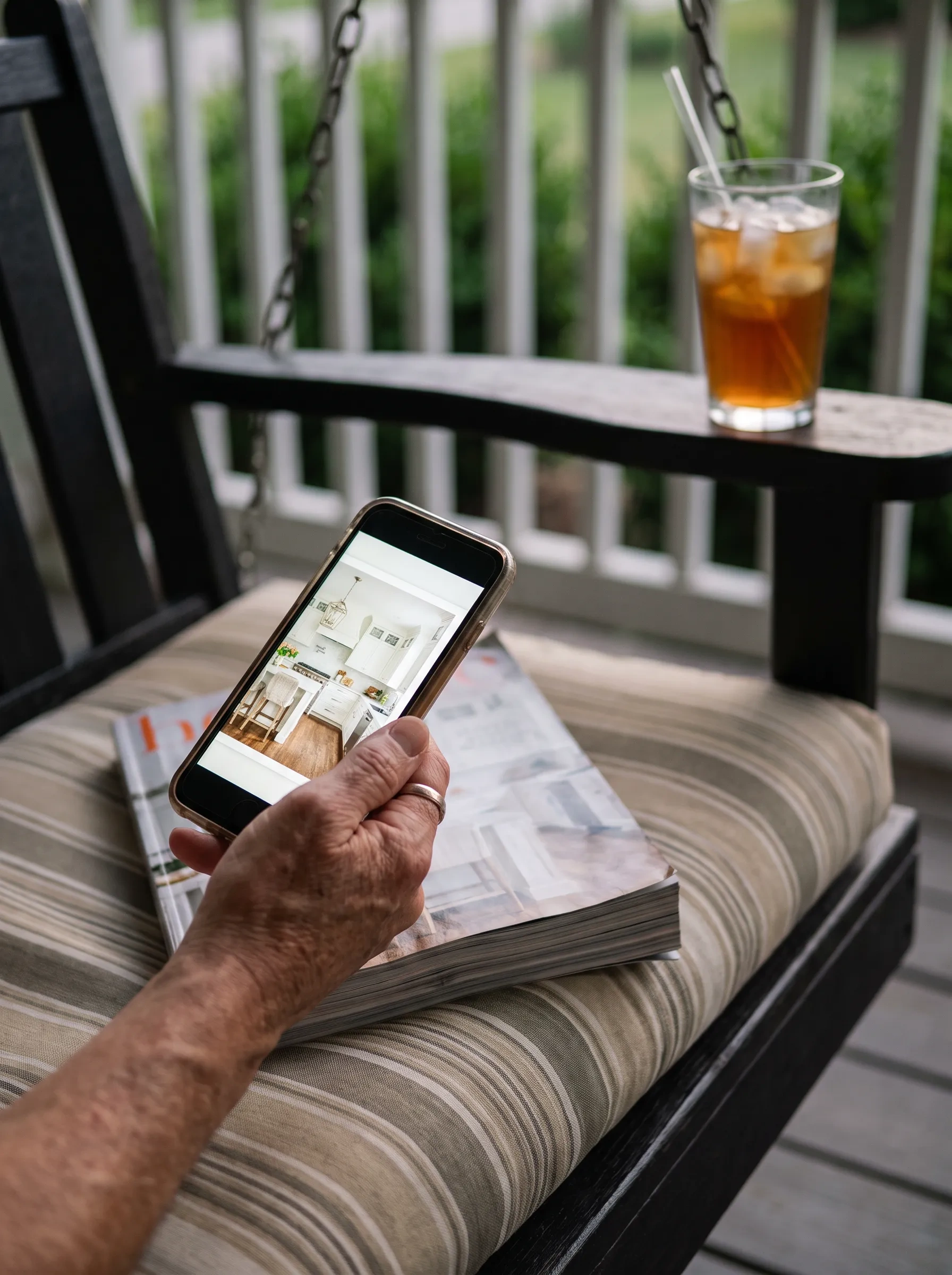 A phone showing a magazine page of a remodeled kitchen on a porch swing, the printed copy on the cushion beside it