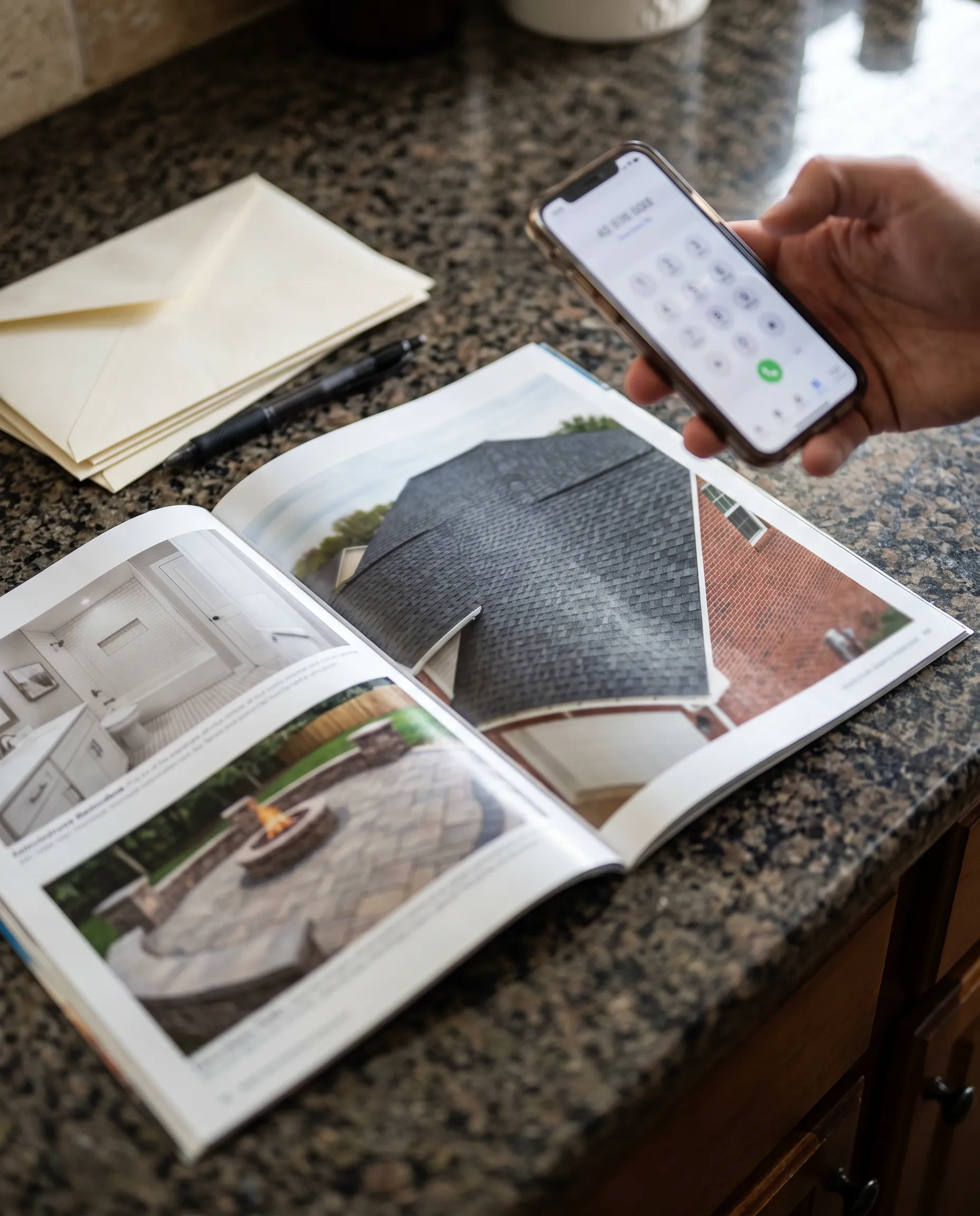 An open home services magazine on a kitchen counter, a roofing ad on the page and a hand holding a phone ready to call