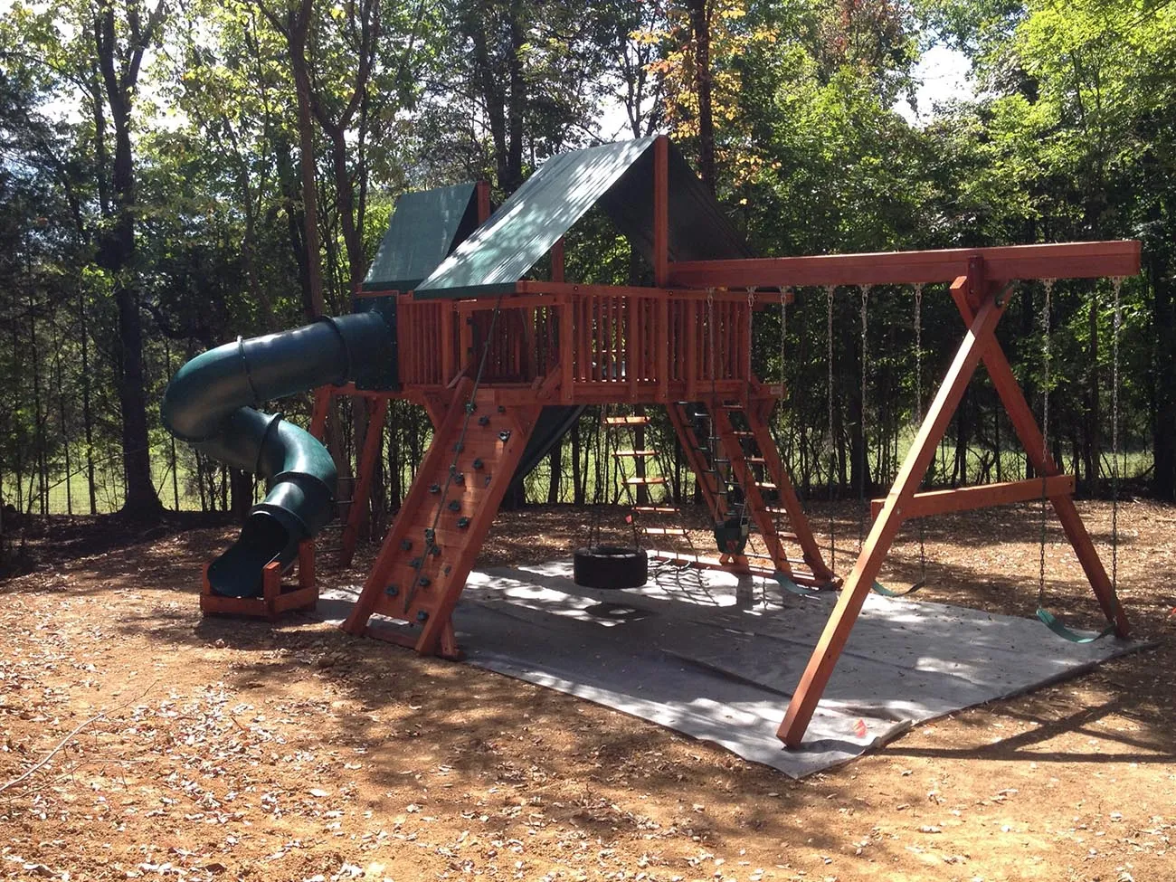 A redwood playset with a green tube slide, a tire swing, a climbing wall and a swing beam, set on a ground cloth at the edge of a wooded lot
